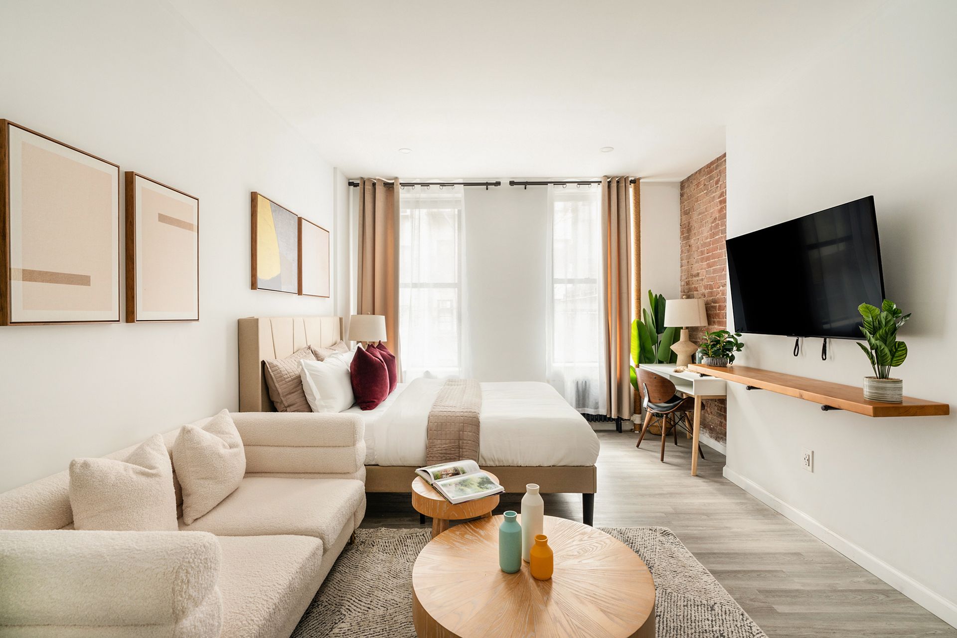 Contemporary studio apartment with a beige sofa, round wooden coffee table, wall-mounted TV, potted plants, large windows with white curtains, and exposed brick walls. The apartment has a washer and dryer and is located in the Upper East Side.
