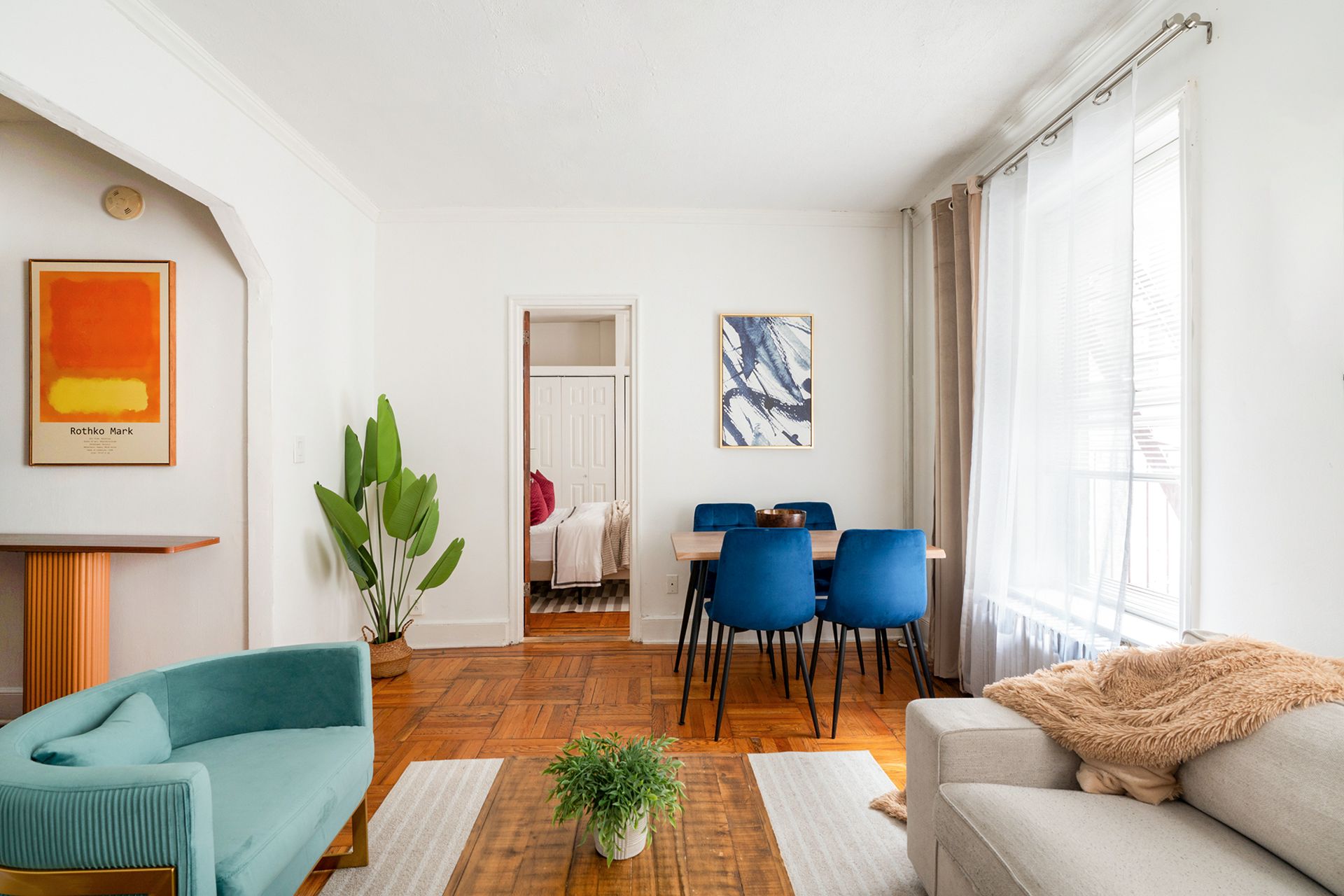 Spacious two-bedroom apartment with a dining area featuring a wooden table, blue chairs, potted plants, and large windows with beige curtains. The apartment is located in Midtown East.