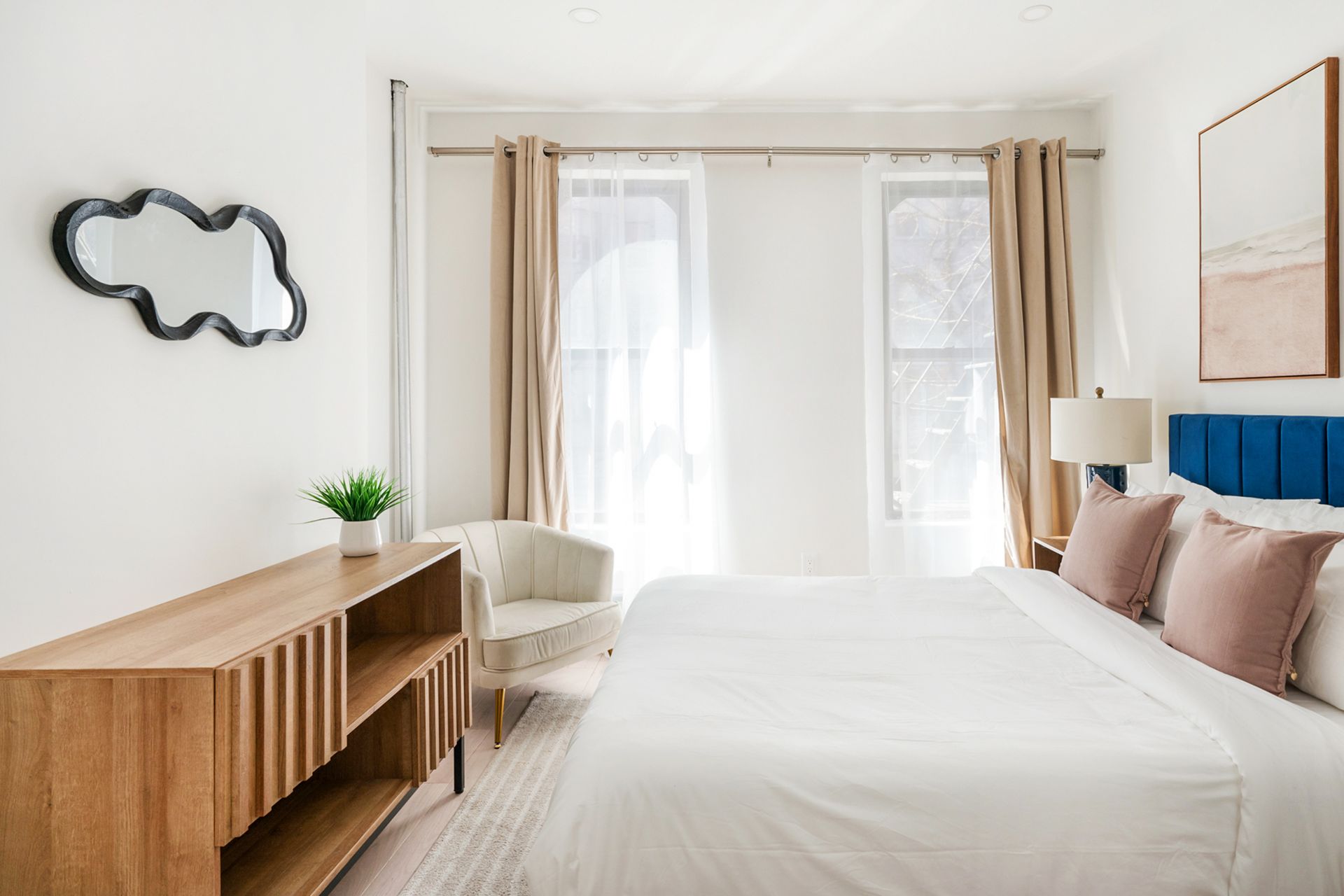 Modern studio apartment with a white bed, blue upholstered headboard, round wall mirror, potted plant, armchair, wooden TV stand, wall-mounted TV, and exposed brick walls. The apartment features large windows with sheer curtains and is located in the Upper East Side.