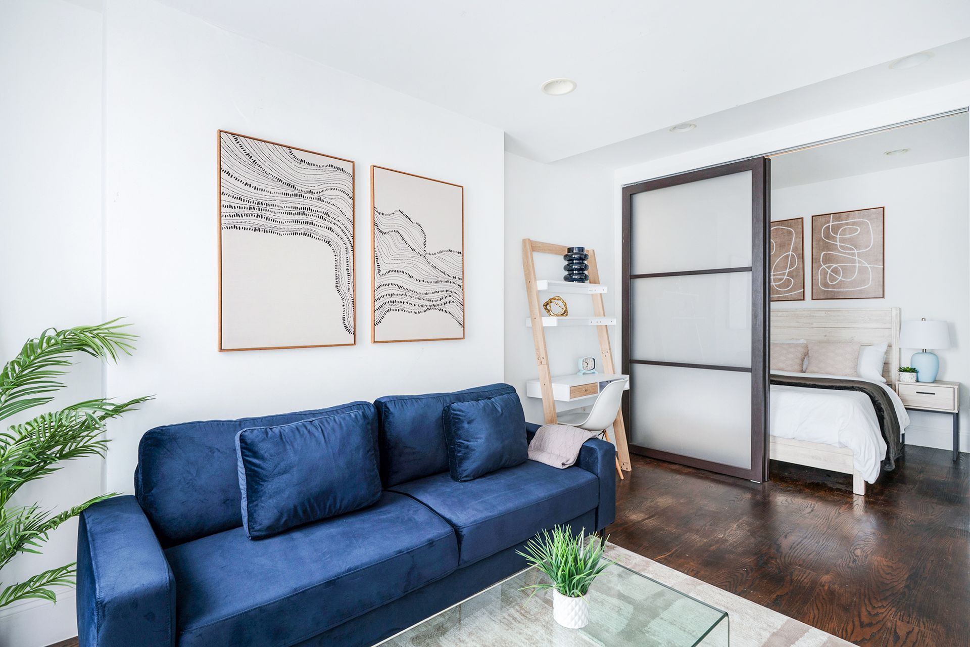 Stylish 1-bedroom apartment in the Lower East Side with a yellow sofa, minimalist wall art, a glass coffee table, a plant, a small desk with a shelf, sliding doors leading to a bright bedroom, and a TV.