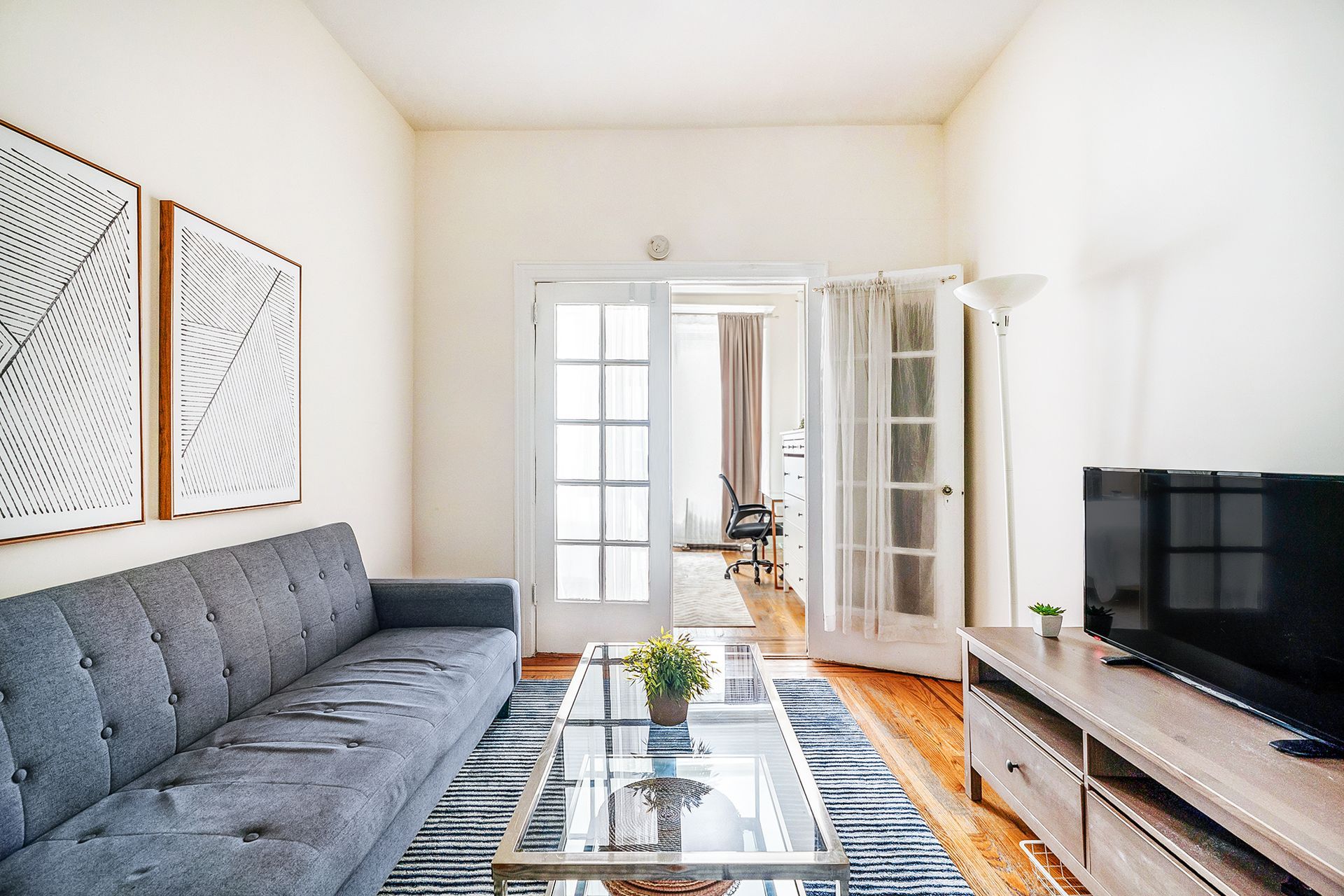 Cozy 1-bedroom apartment in Union Square with a floral-patterned bedspread, a large mirror, a floor lamp, and natural light streaming through sheer curtains, complemented by a green plant.