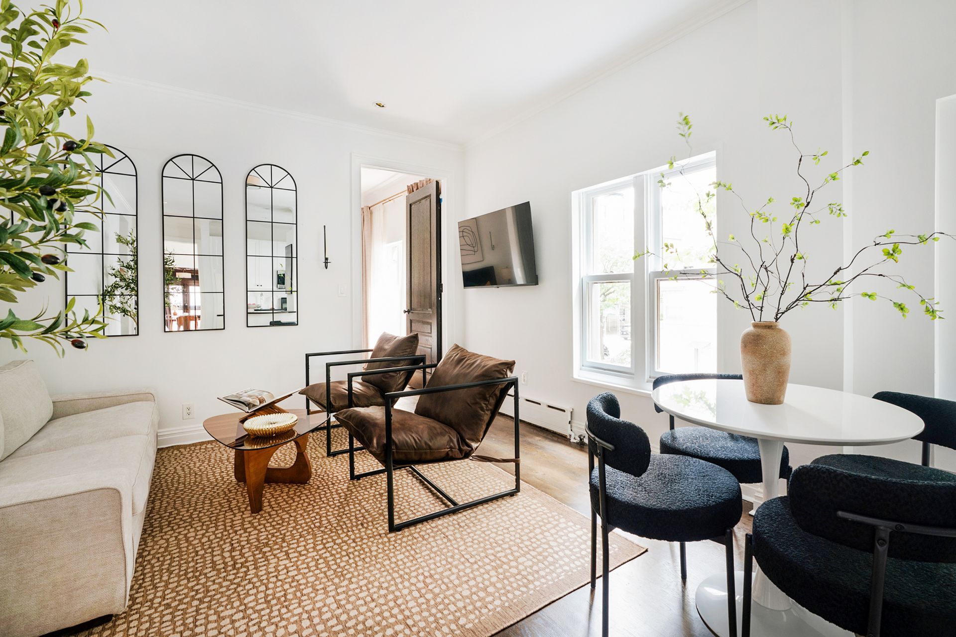 Charming 2-bedroom apartment in the West Village with a cozy and inviting bedroom. The room features a wooden bed frame with white and orange bedding, a tall plant, and a modern bedside table with a lamp. The space is adorned with warm-toned artwork, a leather chair, and a full-length mirror, creating a comfortable and stylish environment.