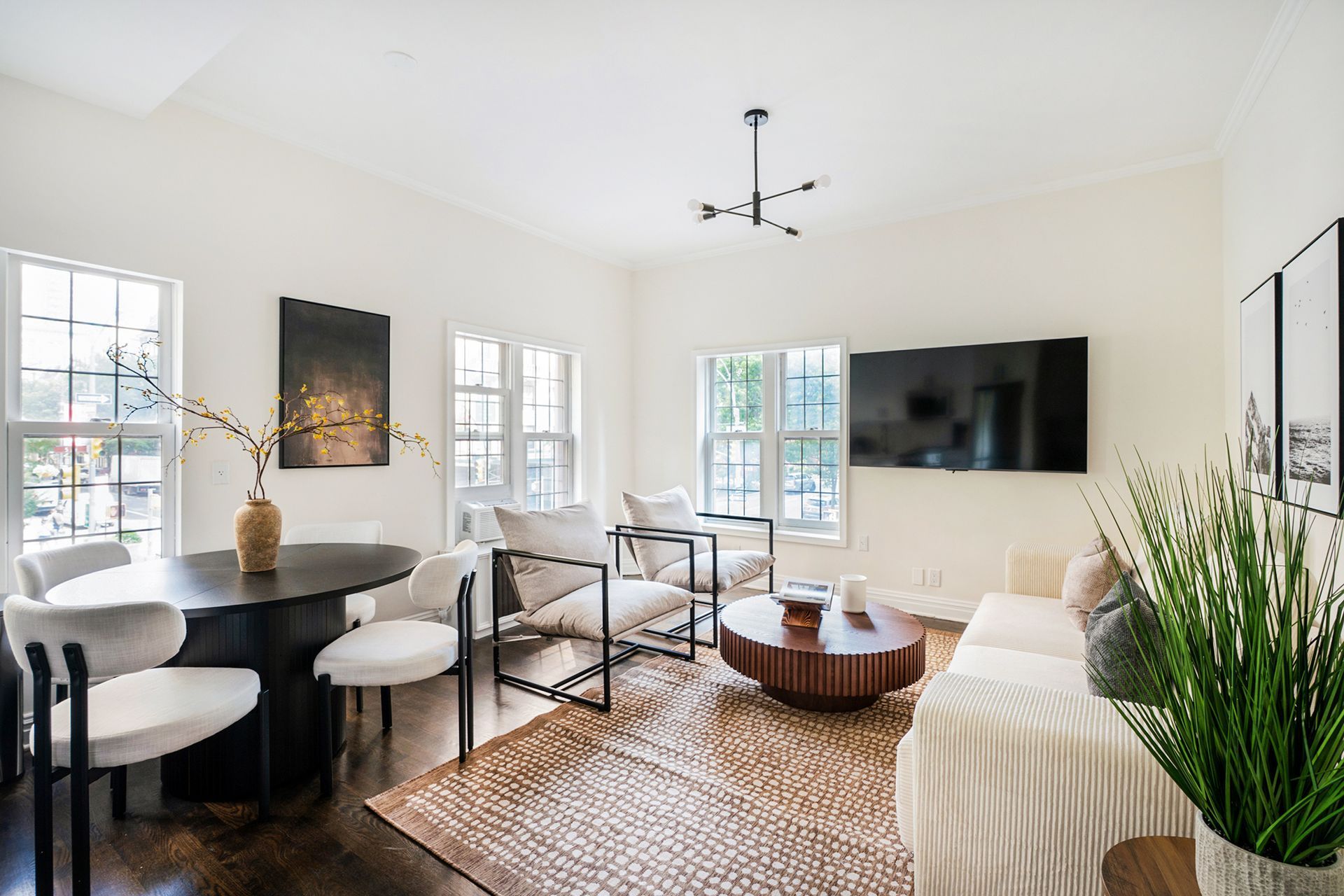 Spacious 2-bedroom apartment in the West Village with a bright and airy bedroom featuring a ceiling fan, large windows with white curtains, and a mix of contemporary and rustic furniture. The room includes a comfortable bed with a wooden frame, accented with red and beige pillows and throws. A modern bedside table with a lamp and potted plant adds a touch of elegance.