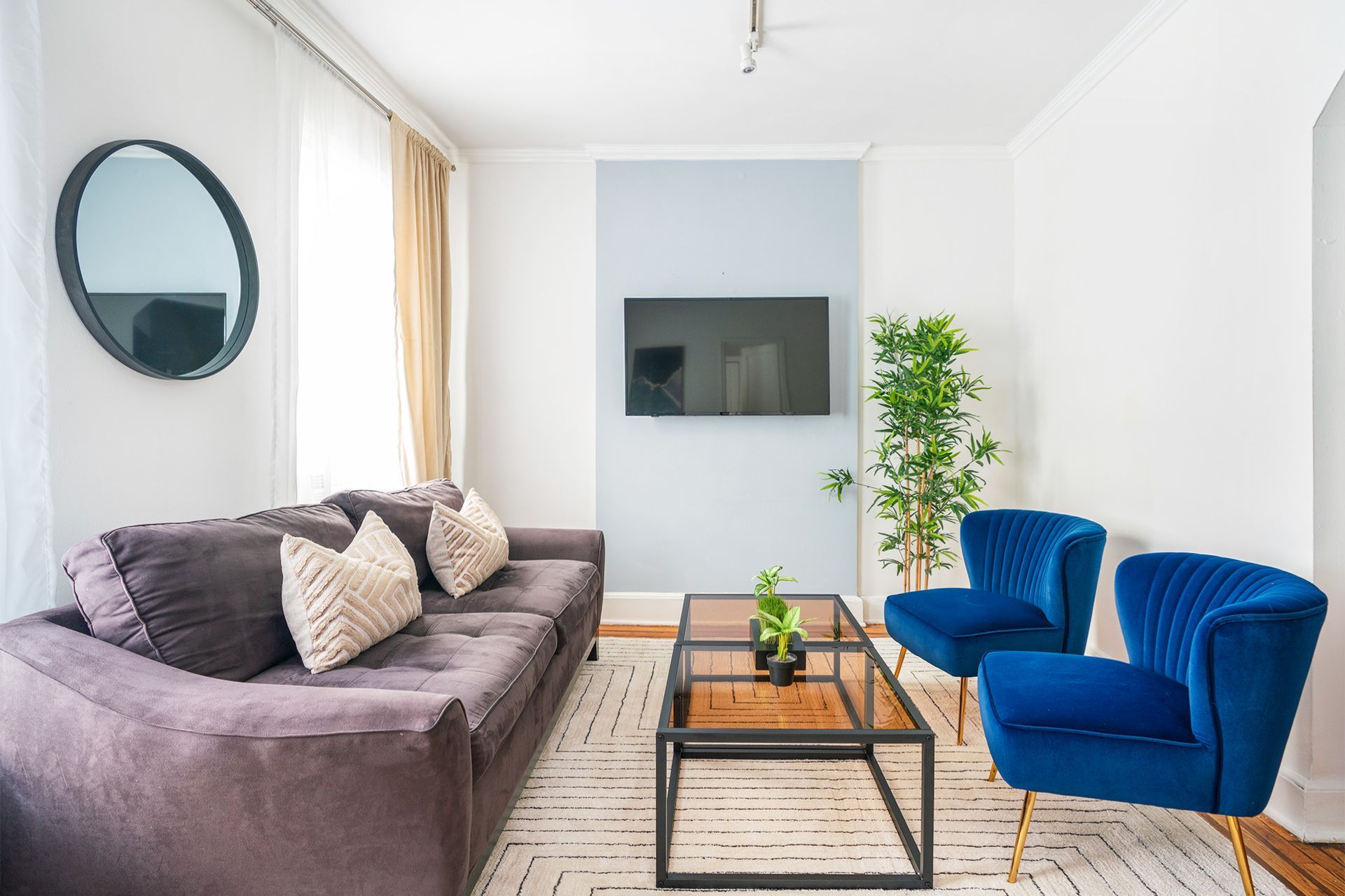 Modern two-bedroom apartment with a gray sofa, blue armchair, wooden coffee table, wall-mounted TV, potted plant, and large windows with beige curtains. The apartment is located in Midtown East.