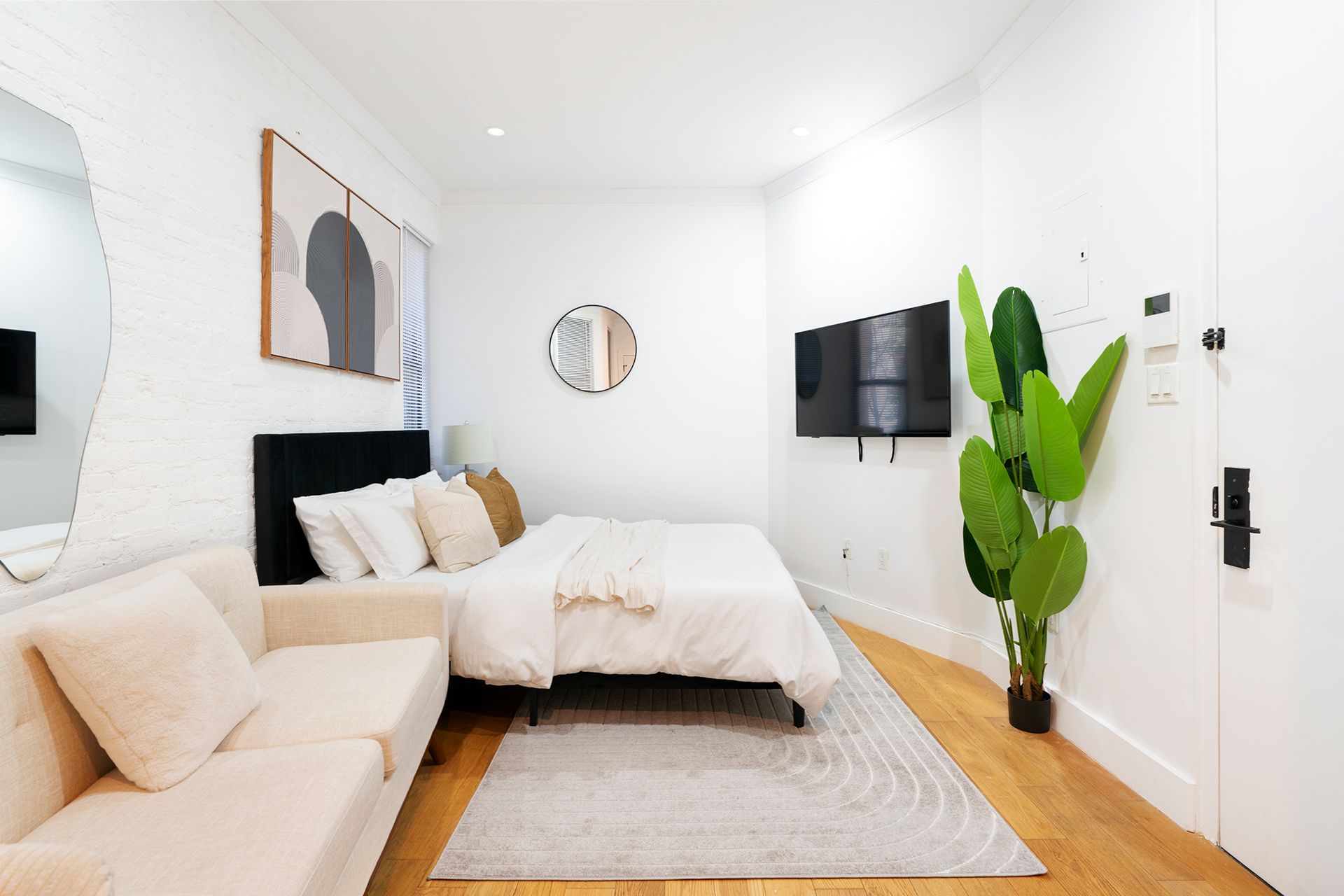 Contemporary studio apartment with a white bed, light beige sofa, wooden coffee table, wall-mounted TV, and potted plants. The apartment has a round wall mirror and is located in the Upper East Side.
