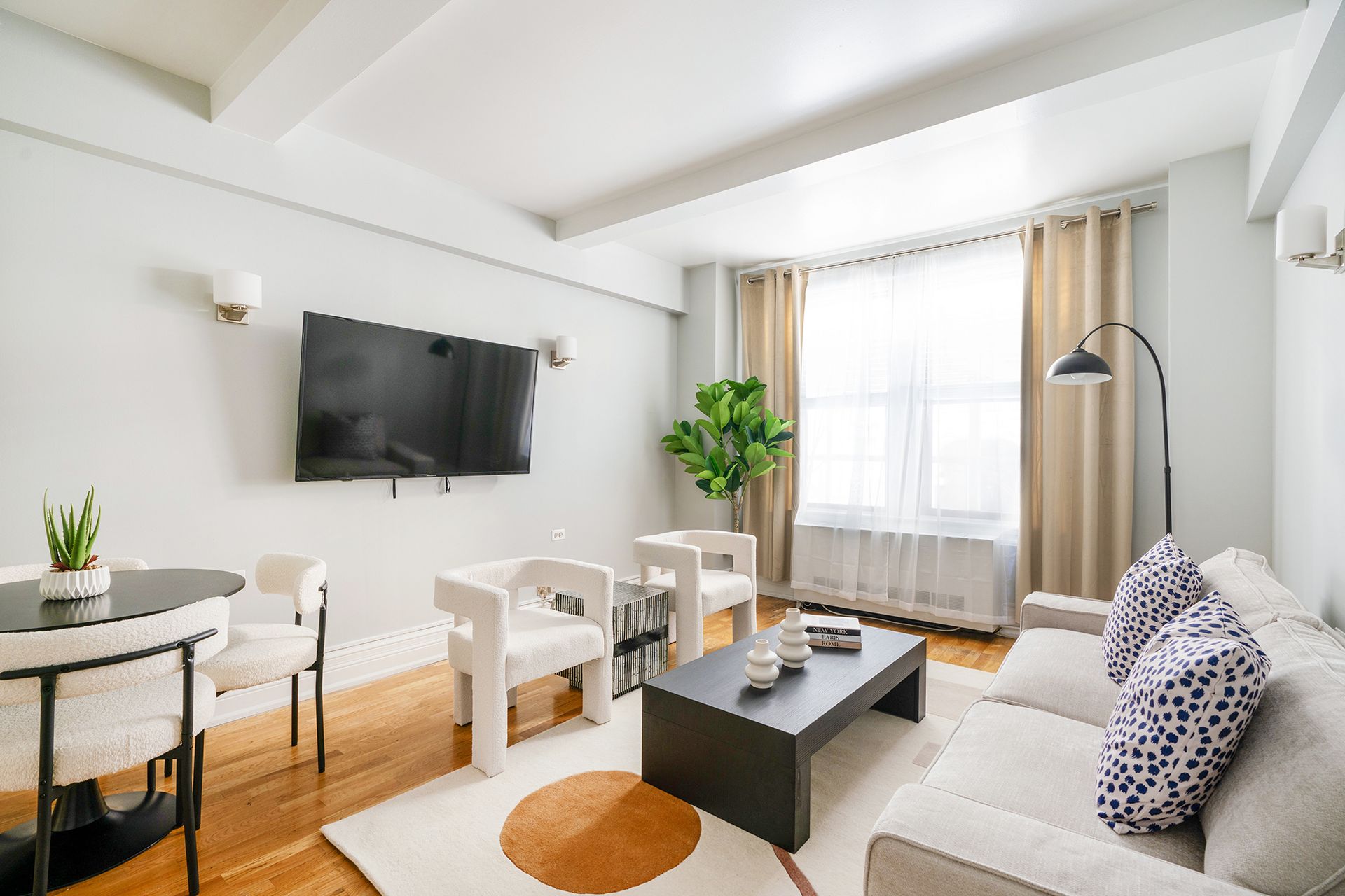 Spacious two-bedroom apartment with a gray sofa, wooden coffee table, dining area with white chairs, wall-mounted TV, and large windows with beige curtains. The apartment includes a doorman and gym and is located in the Upper West Side.