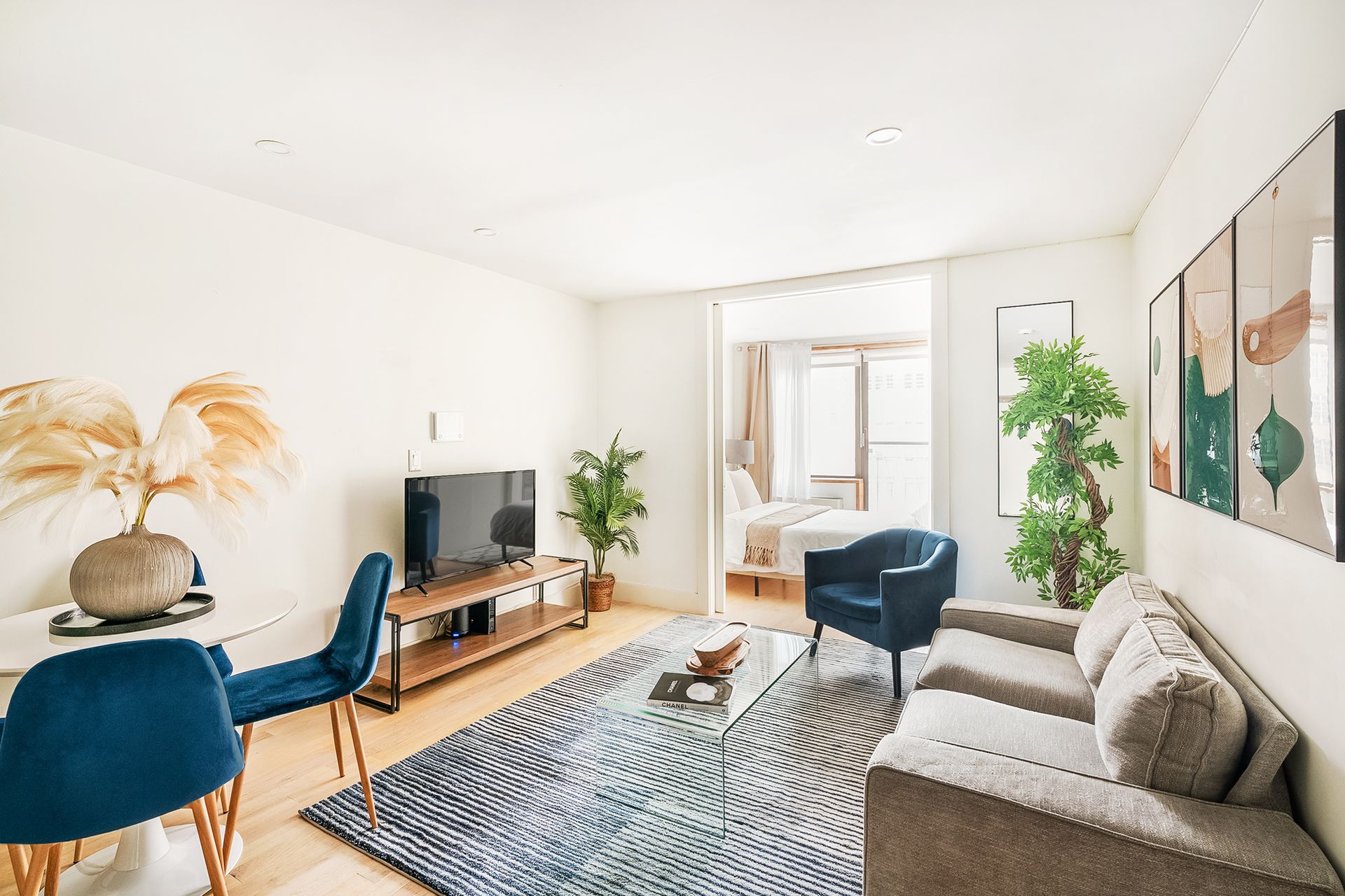 Bright three-bedroom apartment with a yellow sofa, glass coffee table, dining area with a bar-height table and stools, modern kitchen with stainless steel appliances, and a glass staircase. The apartment has a washer and dryer.