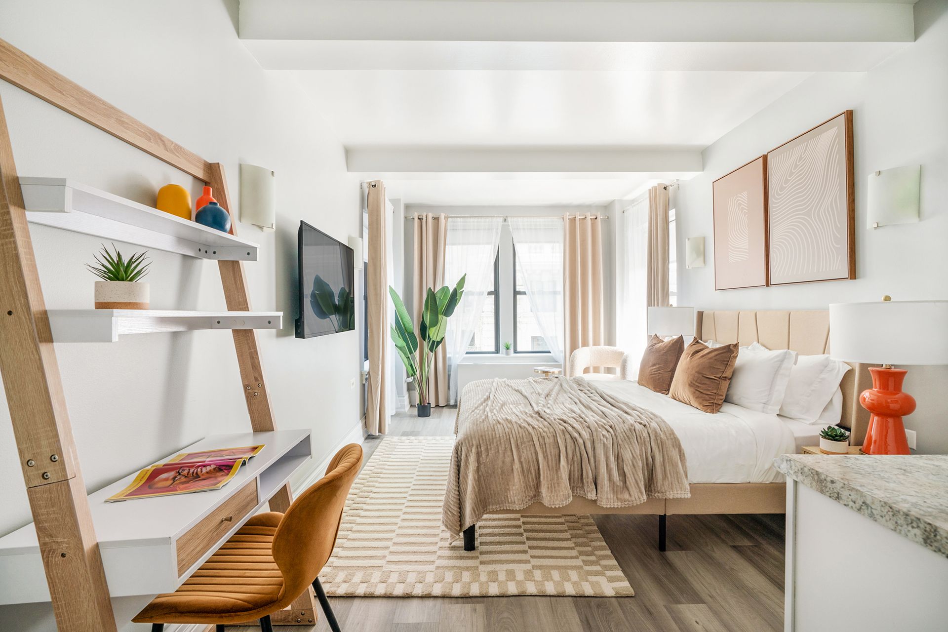 Modern studio apartment with a white bed, beige throw blanket, wooden desk with an orange chair, wall-mounted TV, potted plant, and large windows with beige curtains. The apartment includes a doorman, gym, and laundry facilities and is located in the Upper West Side.