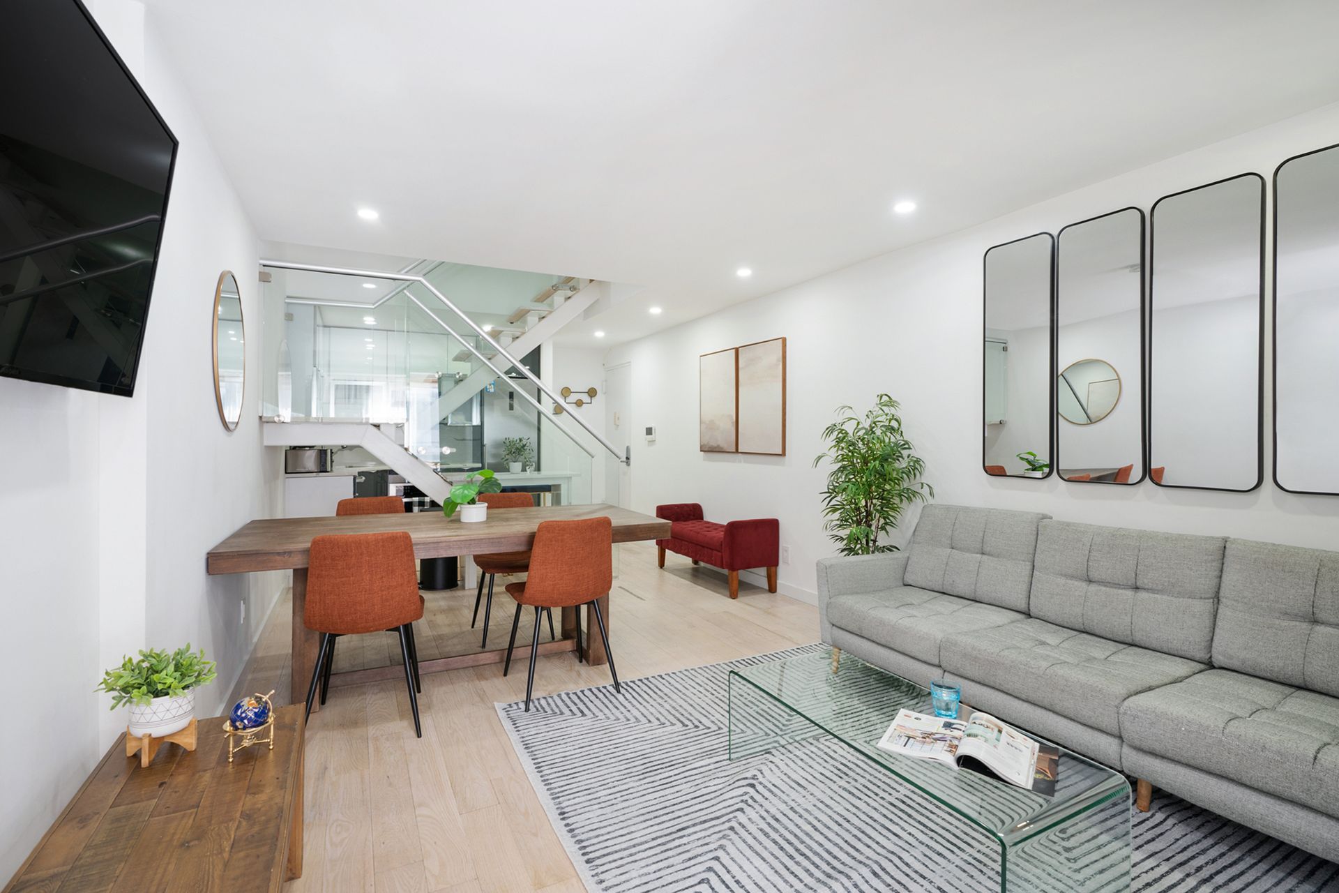 Contemporary three-bedroom apartment with a gray sofa, glass coffee table, dining area with orange chairs, modern kitchen, and glass staircase. The apartment has a washer and dryer and an in-unit elevator.