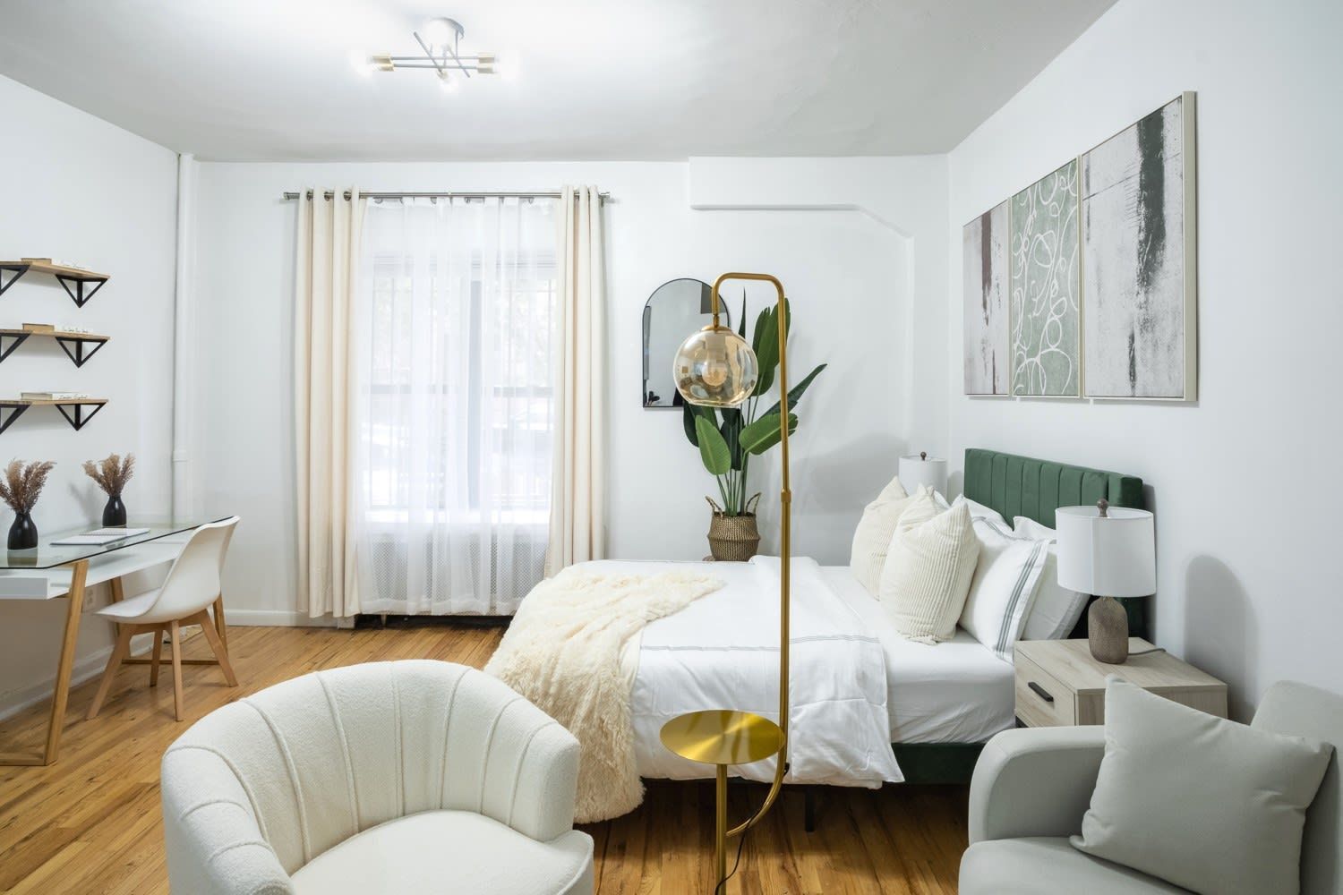 Bright studio apartment with a white bed, beige throw blanket, green upholstered headboard, white armchair, wooden desk with a chair, potted plant, and large windows with beige curtains. The apartment includes access to a gym and is located in the Upper West Side.