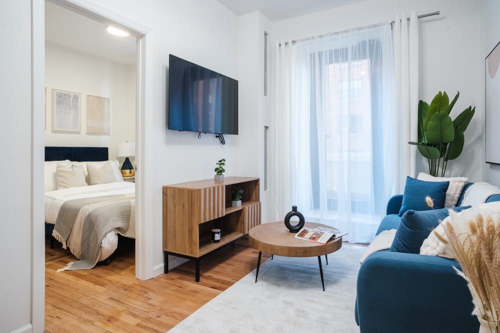 Spacious two-bedroom apartment with a blue sofa, wooden coffee table, wall-mounted TV, white bed, potted plant, and large windows with sheer curtains. The apartment includes a washer and dryer, access to a gym, and is located in the Upper West Side