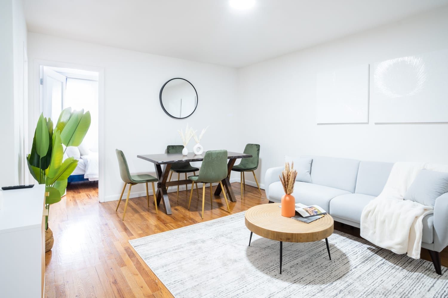 Modern two-bedroom apartment with a white sofa, gray dining chairs, round wooden dining table, round wall mirror, potted plant, and large windows. The apartment includes a washer and dryer and is located in Midtown East.