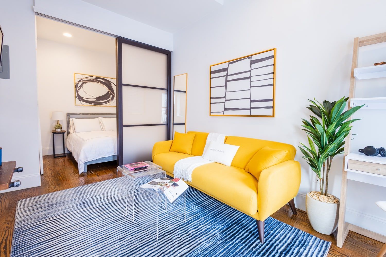 Contemporary 1-bedroom apartment in the Lower East Side featuring a yellow sofa, decorative wall art, a glass coffee table, a cozy workspace with a shelf, a plant, and sliding doors leading to a modern bedroom.