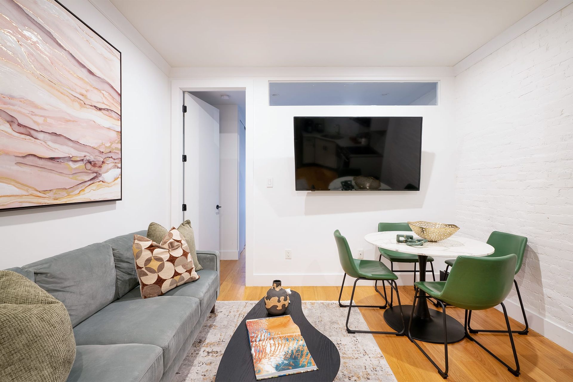Modern two-bedroom apartment with a gray sofa, black coffee table, wall-mounted TV, green dining chairs, and a colorful rug. The apartment is located in the Upper East Side.