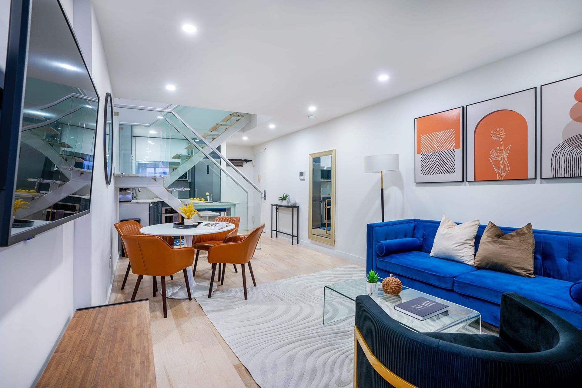 Contemporary three-bedroom apartment with a blue sofa, orange chairs, glass coffee table, dining area, modern kitchen with white cabinets, and wall art. The apartment has a washer and dryer.