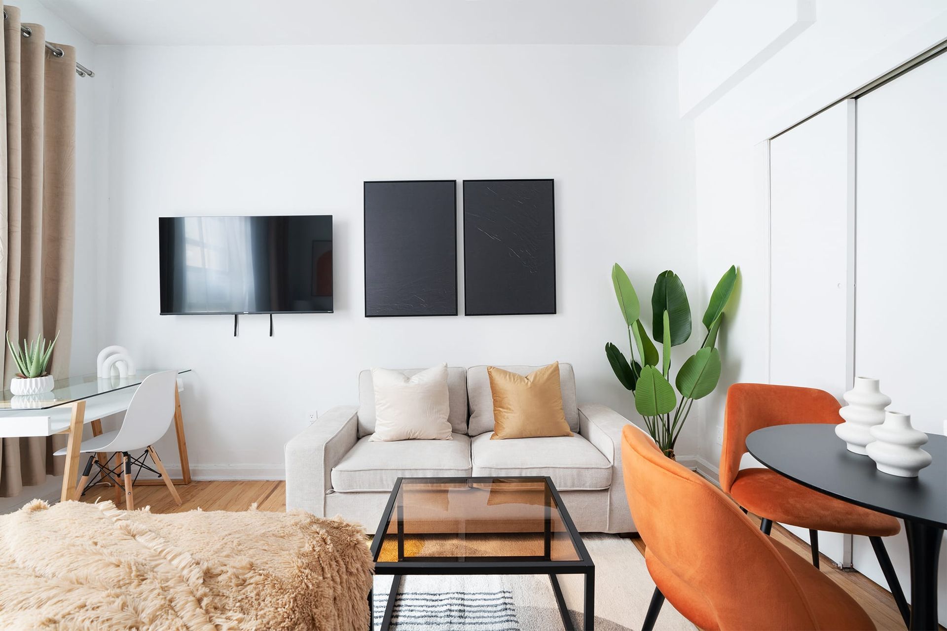 Contemporary studio apartment with a beige sofa, orange dining chairs, black coffee table, wall-mounted TV, potted plant, and large windows with beige curtains. The apartment includes access to a gym and is located in the Upper West Side.