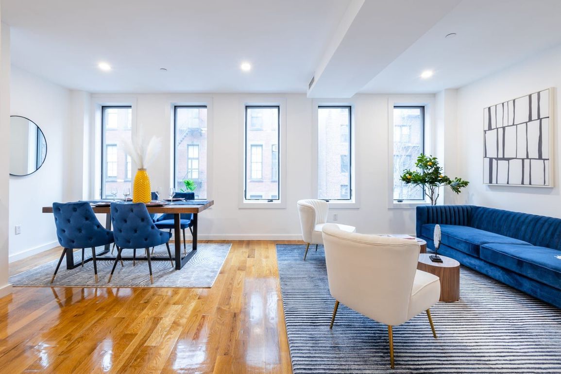 Bright, modern living space with wooden floors, blue velvet sofa, dining table, and large windows.
