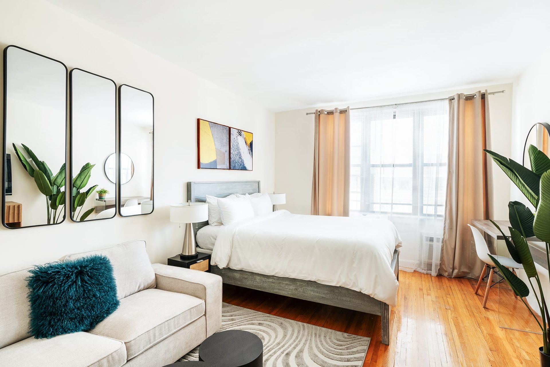 Bright and airy Studio apartment in Union Square featuring a modern bedroom with a white bedspread, large windows with beige curtains, a wall-mounted mirror, indoor plants, and a small desk area.
