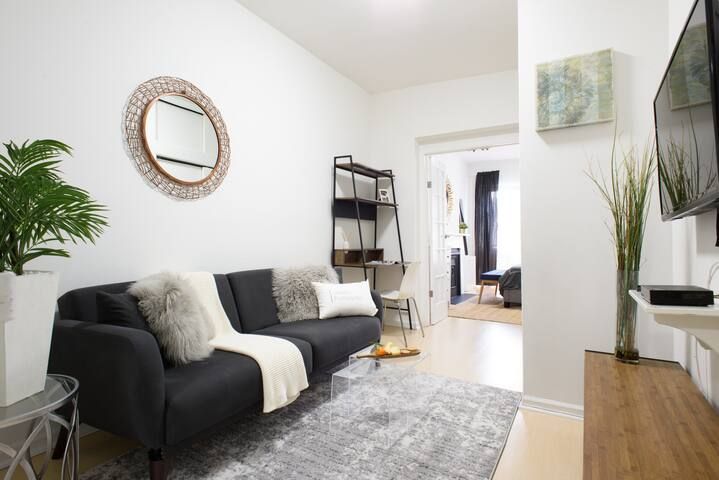 Contemporary two-bedroom apartment with a dark gray sofa, gray area rug, decorative wall mirror, potted plant, and a wall-mounted TV. The apartment has wooden floors and is located in the Upper East Side.