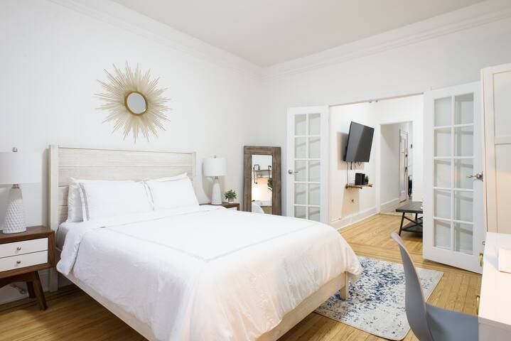 Bright two-bedroom apartment with a white bed, sunburst wall mirror, bedside tables with lamps, large standing mirror, and French doors leading to a living area with a wall-mounted TV. The apartment includes a washer and dryer and is located in the Upper East Side.