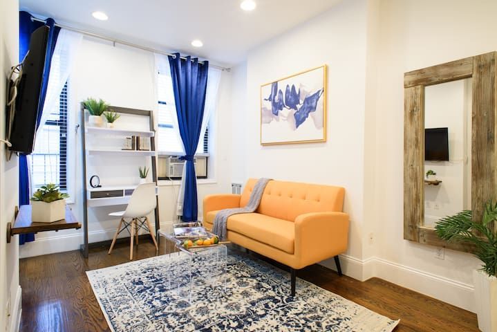 Modern 1-bedroom apartment in the Lower East Side with a vibrant yellow sofa, blue area rug, wall-mounted art, blue curtains, and a wall mirror, creating a bright and welcoming space.