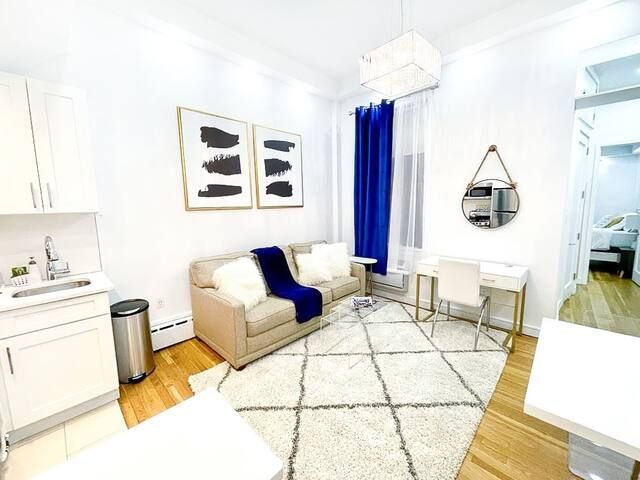 Bright one-bedroom apartment with a beige sofa, white armchair, wooden coffee table, large wall mirror, and large windows with blue curtains. The apartment is located in Midtown East.