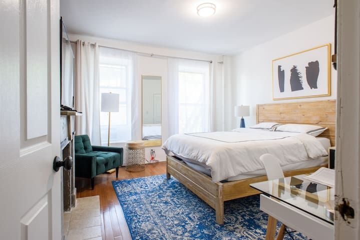 Spacious two-bedroom apartment with a cozy bedroom featuring a wooden bed frame, white bedding, a blue area rug, and large windows allowing natural light. Located in Midtown West.