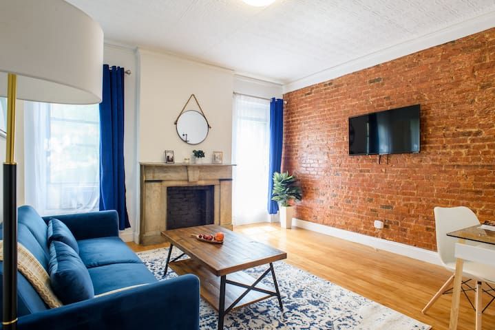 2 bedroom apartment with living room that includes sofa, coffee table, brick wall, fireplace, and TV. Located in Midtown West
