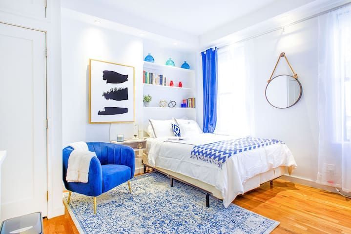 Contemporary studio apartment with a white bed, blue armchair, clear coffee table, wooden desk with a chair, wall-mounted TV, potted plant, and large windows with blue curtains. The apartment includes a washer and dryer and is located in Midtown East.