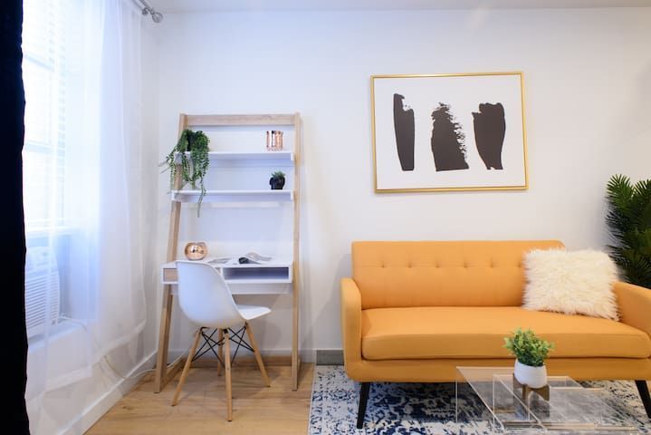Charming 1-bedroom apartment in the Lower East Side with a yellow sofa, minimalist wall art, a blue area rug, a small workspace with a ladder shelf, and a glass coffee table.