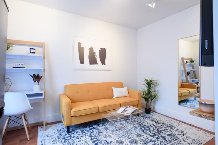 Charming 1-bedroom apartment in the Lower East Side with a yellow sofa, minimalist wall art, a glass coffee table, a small workspace with a shelf, a plant, and a large mirror reflecting the bright, airy space.