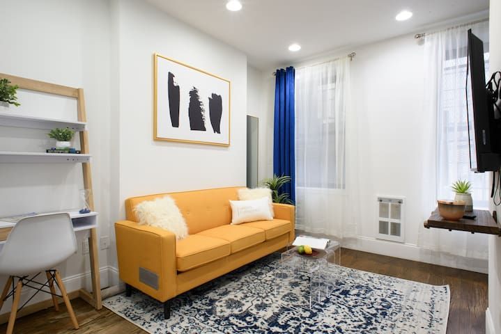 Cozy 1-bedroom apartment in the Lower East Side featuring a bright living room with a yellow sofa, white walls, a decorative wall art, blue curtains, and a small workspace with a ladder shelf.