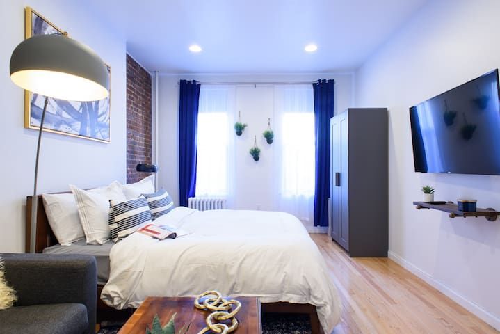 Modern studio apartment with a white bed, gray armchair, wooden coffee table, wall-mounted TV, large windows with blue curtains, and a floor lamp. The apartment features exposed brick walls and is located in the Upper East Side.