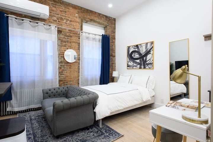Modern one-bedroom apartment with a white bed, gray armchair, wooden coffee table, wall-mounted TV, large wall mirror, and exposed brick walls. The apartment includes a washer and dryer and is located in Midtown East.