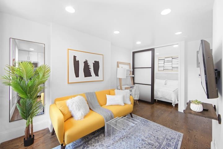 Stylish 1-bedroom apartment in the Lower East Side with a yellow sofa, a blue area rug, minimalist wall art, a glass coffee table, a cozy workspace with a ladder shelf, sliding doors leading to the bedroom, and a TV.
