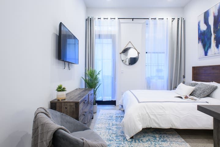 Contemporary 1-bedroom apartment in the East Village showcasing a neatly arranged bedroom with a wooden headboard and crisp white bedding. The room has a large window with sheer curtains, a mounted flat-screen TV, and a rustic wooden dresser. The space is decorated with a round mirror, a small plant, and modern artwork, creating a stylish and comfortable atmosphere.