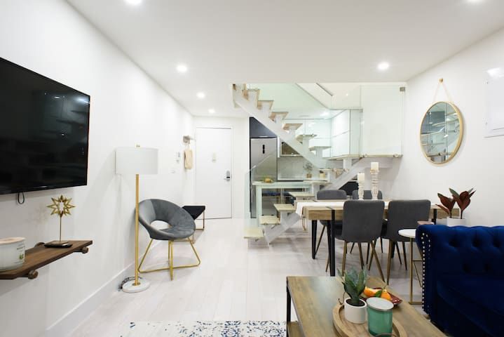Spacious three-bedroom apartment with a dining area, glass dining table, black chairs, modern kitchen with white cabinets, and a TV mounted on the wall. The apartment has a washer and dryer.