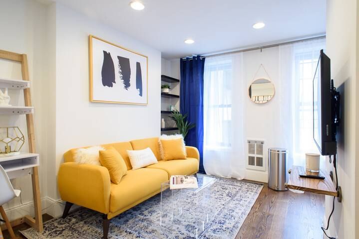 Bright and modern 1-bedroom apartment in the Lower East Side featuring a vibrant yellow sofa, a blue area rug, minimalist wall art, blue curtains, a glass coffee table, a small desk, and a TV.