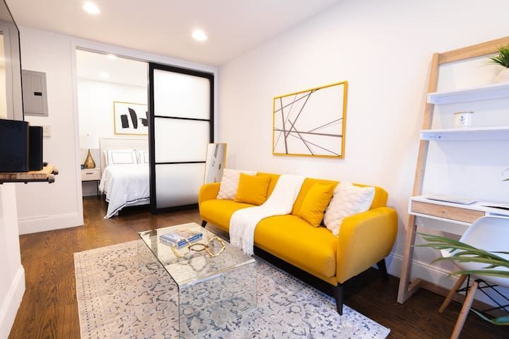 Contemporary 1-bedroom apartment in the Lower East Side featuring a yellow sofa, decorative wall art, a glass coffee table, a cozy workspace, and sliding doors leading to the bedroom.