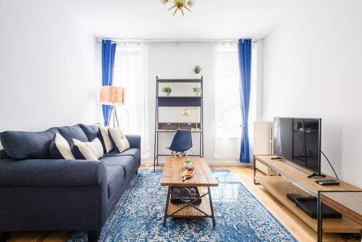 2-bedroom apartment with living room, sofa, coffee table, TV stand, and  window with blue curtains. Located in Midtown West.