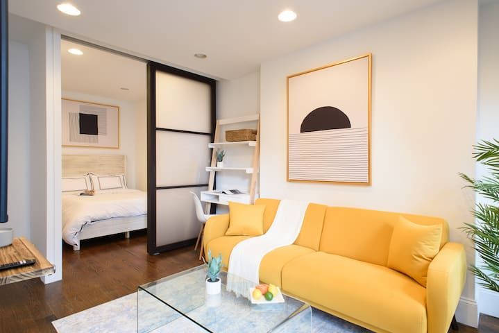 Modern 1-bedroom apartment in the Lower East Side featuring a yellow sofa, minimalist wall art, a glass coffee table, a small desk with a shelf, sliding doors leading to a cozy bedroom, and a TV.