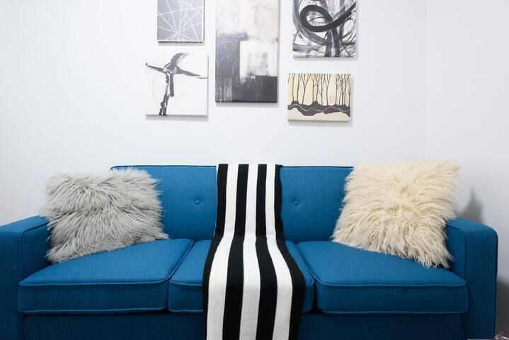 Modern two-bedroom apartment with a blue sofa, fluffy gray and white throw pillows, a black and white striped blanket, and abstract wall art. The apartment includes a dishwasher and dryer and is located in the Upper East Side.