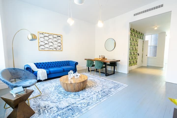 Elegant 1-bedroom designer townhouse in the East Village showcasing a spacious living and dining area. The room is decorated with a plush blue couch, a round wooden coffee table, and a dining table with chairs. The light walls contrast beautifully with the blue accents, and the space is complemented by a large mirror, modern lighting, and decorative artwork, creating a welcoming and stylish atmosphere.