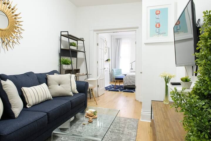 Cozy two-bedroom apartment with a dark blue sofa, clear coffee table, potted plant, wall-mounted TV, and a bookshelf. The apartment has wooden floors and is located in the Upper East Side.