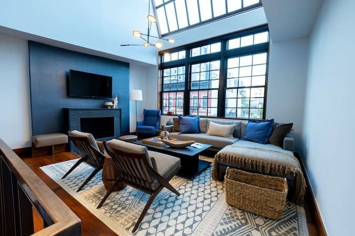 Luxurious 3-bedroom designer townhouse in the East Village, featuring an expansive living room with floor-to-ceiling windows. The space is adorned with comfortable seating including a large sectional sofa, armchairs, and a coffee table on a patterned rug. The high ceilings and ample natural light enhance the room's elegance, and the decor includes a modern fireplace, contemporary lighting fixtures, and tasteful decorative elements.