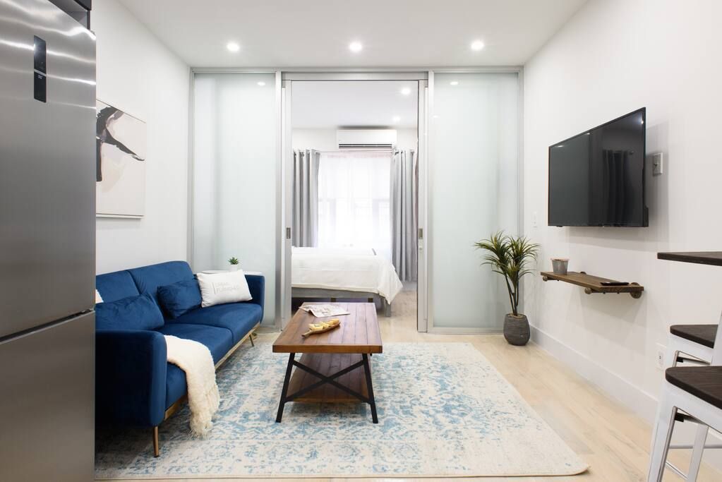 Contemporary one-bedroom apartment with a blue sofa, wooden coffee table, white bed, blue patterned rug, and large frosted glass doors. The apartment has a washer and dryer.