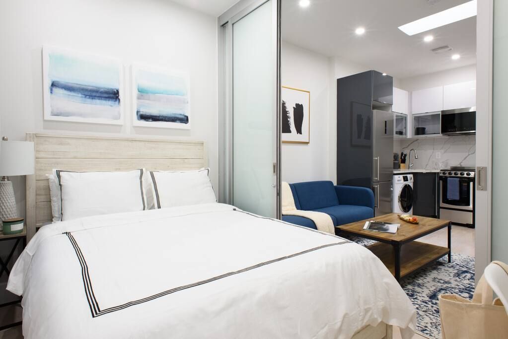 Contemporary one-bedroom apartment with a white bed, blue sofa, wooden coffee table, and large frosted glass doors. The apartment has a washer and dryer and a skylight.