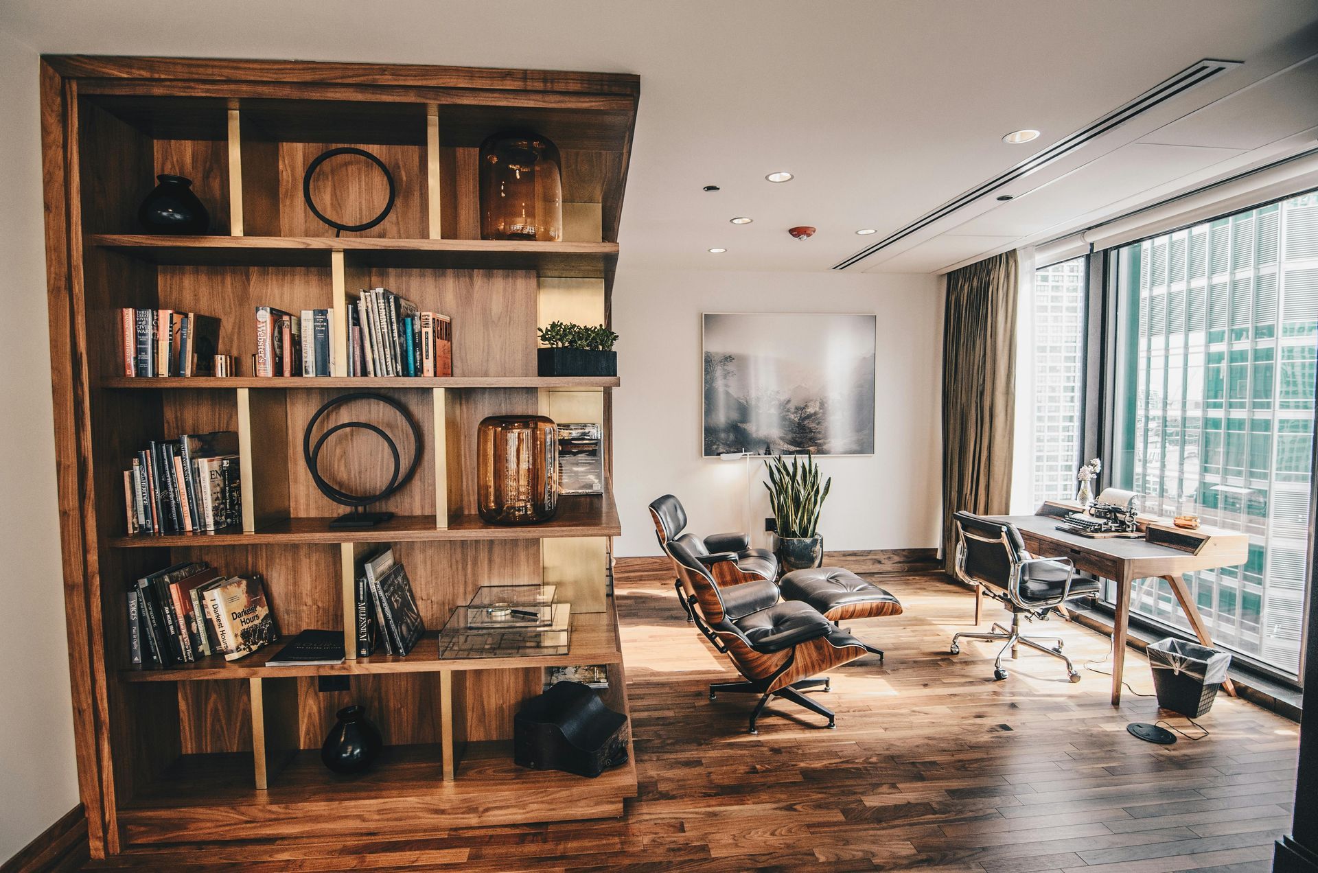 Wooden bookshelf and desk in a modern office with large windows, leather chair, artwork, and hardwood floors.