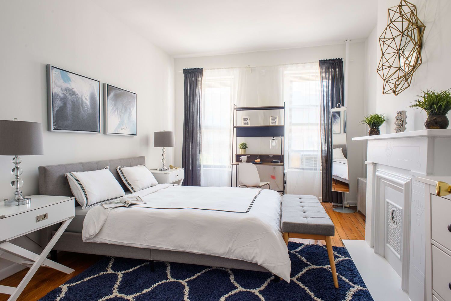 Bright two-bedroom apartment with a white bed, blue patterned rug, large windows with blue curtains, a white fireplace, and a bookshelf. The apartment is located in the Upper East Side.