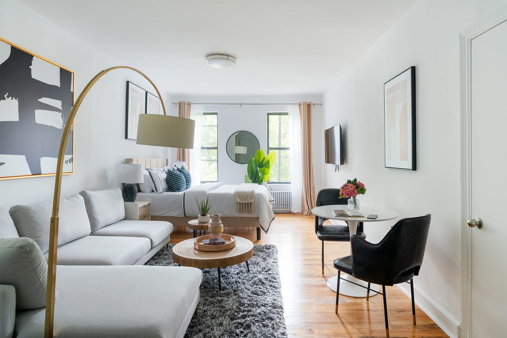 Contemporary studio apartment featuring a cozy seating area with modern furniture, a round coffee table, a comfortable bed with white bedding, and large windows allowing natural light to fill the space. Located in Midtown West.